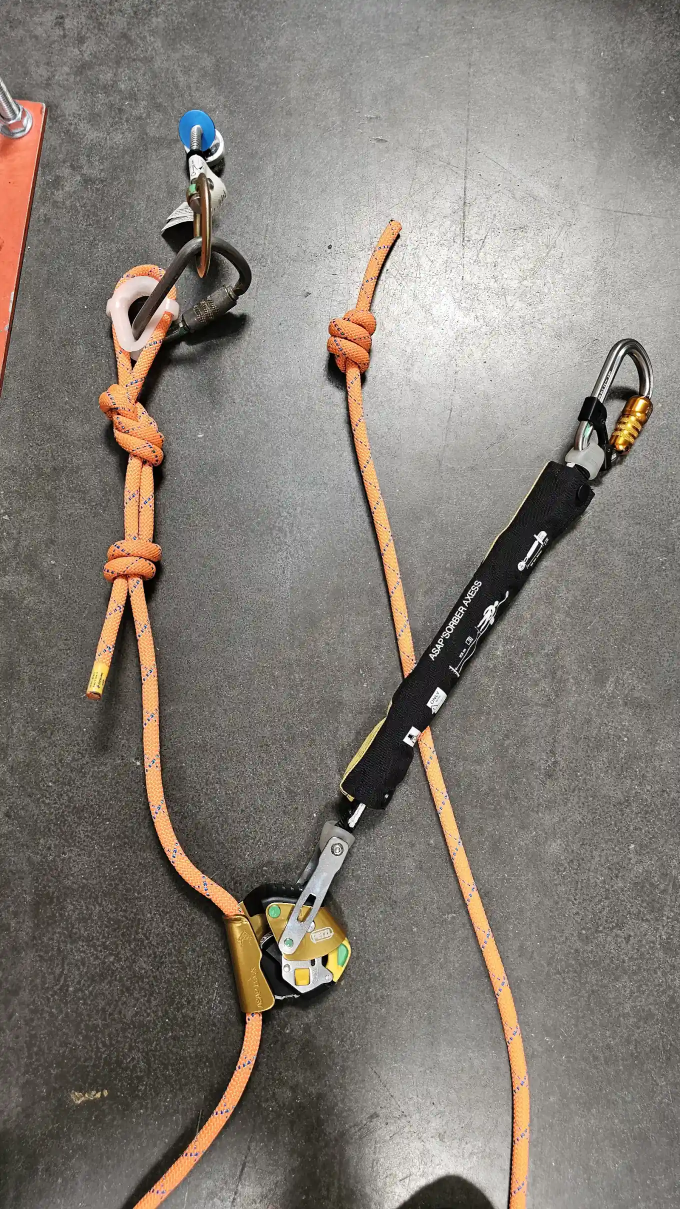 An orange kernmantle rope on a shop floor with a series of knots tied along it, a Petzl ASAP arrester on the line and an ASAP'SORBER AXESS absorber running to a locking carabiner