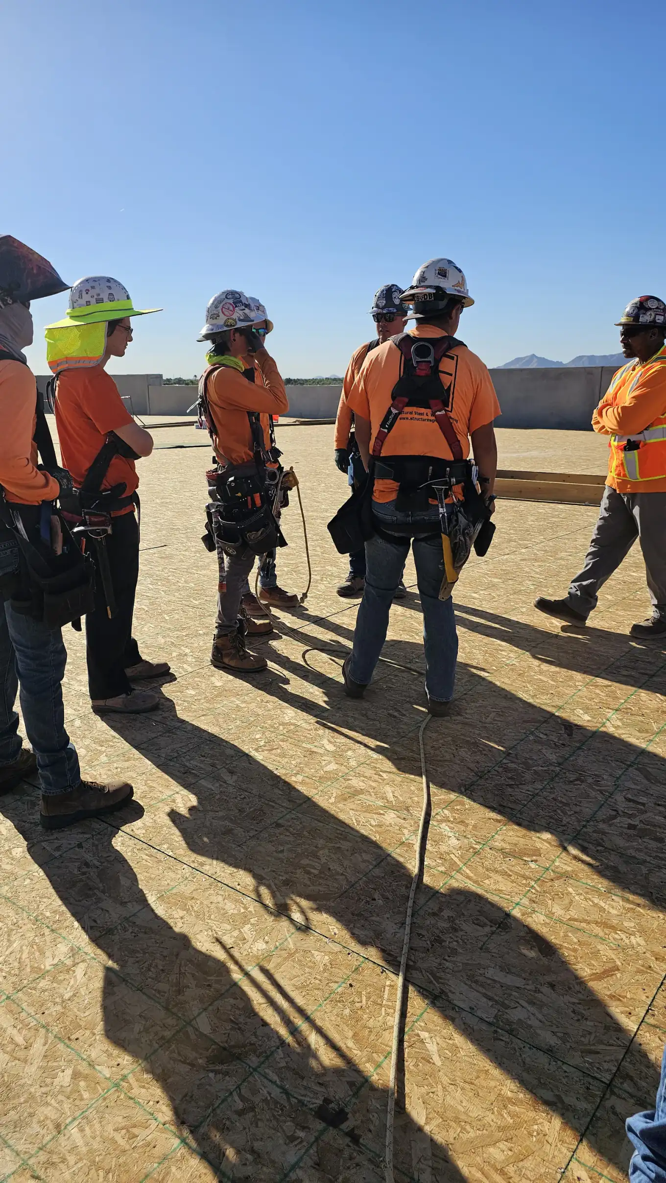 A crew in hard hats, high-visibility shirts and fall protection harnesses standing on a plywood deck at a jobsite during a training session