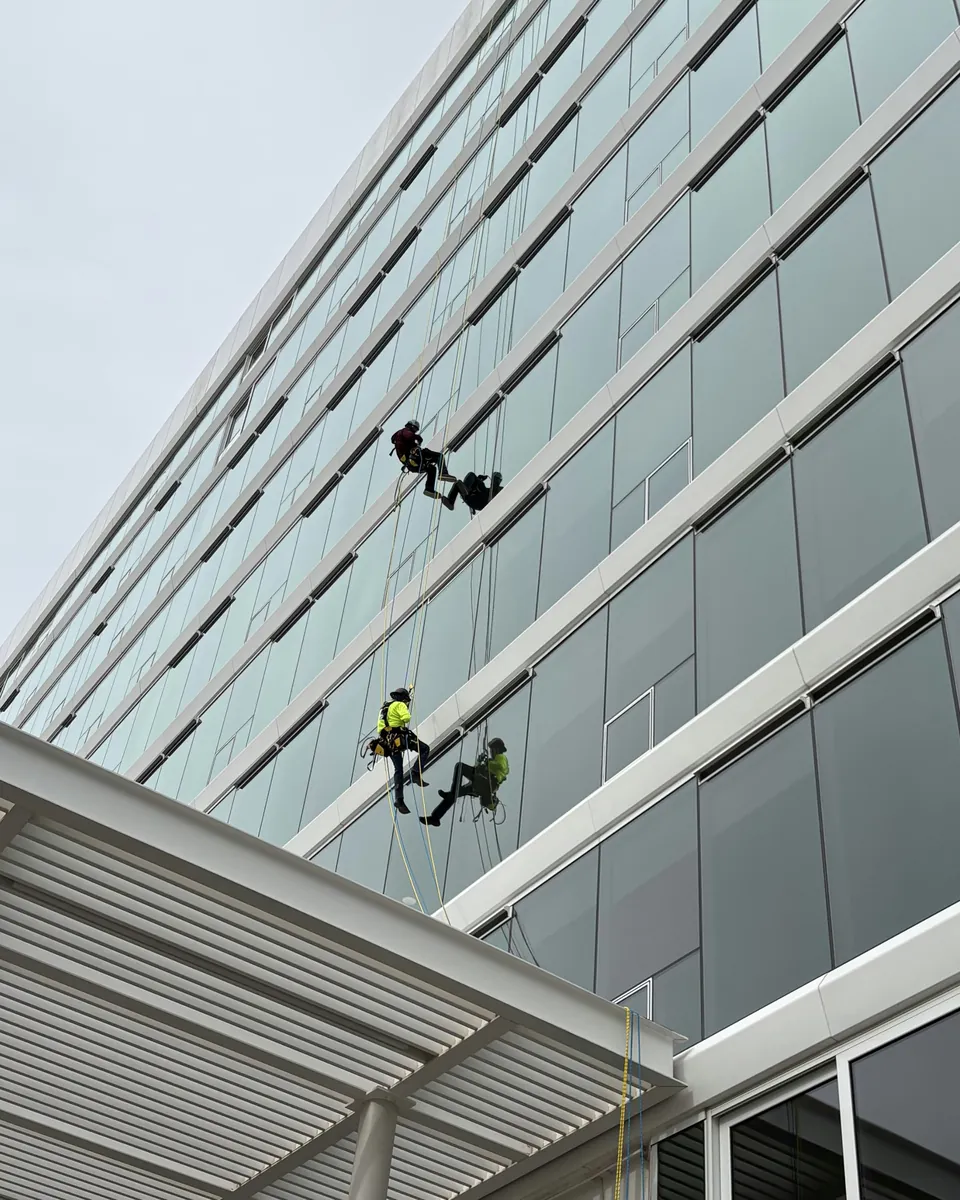 Two workers in helmets and harnesses suspended on ropes against the glass facade of a multistory building, each reflected in the glass beside them
