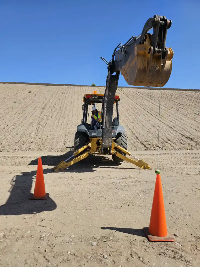 A backhoe operator lowering the bucket toward a traffic cone during a precision skill challenge