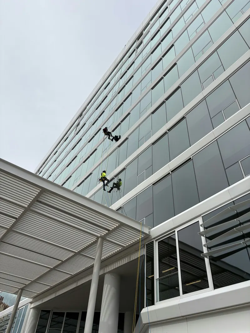 Two rope access workers in helmets and harnesses suspended against the glass facade of a multistory building, each reflected in the glass, above an entrance canopy