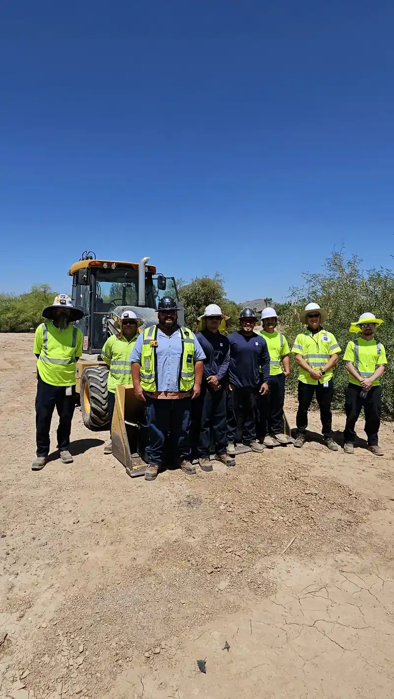 A group of trainees in high-visibility vests in front of a backhoe after heavy equipment training
