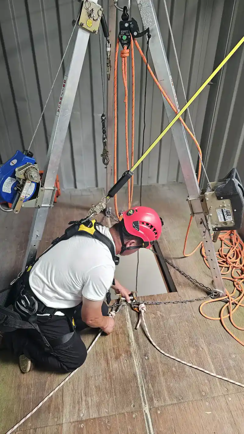 A trainee in a red helmet working at a floor hatch under a rope rescue tripod during confined space entry training