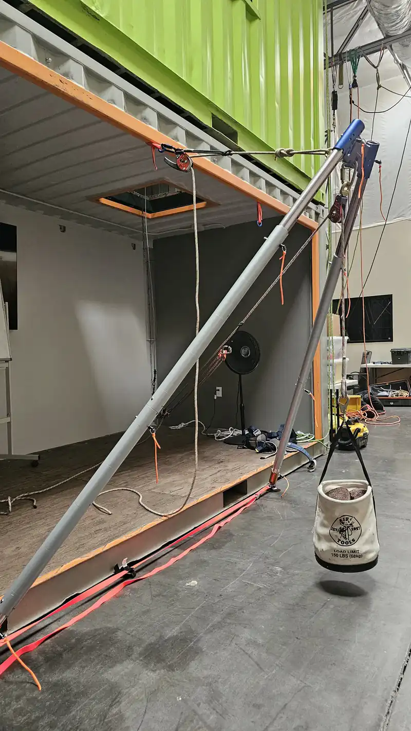 A rigging pole anchored inside an open shipping-container training bay, with a rope bag suspended below and a green container stacked above